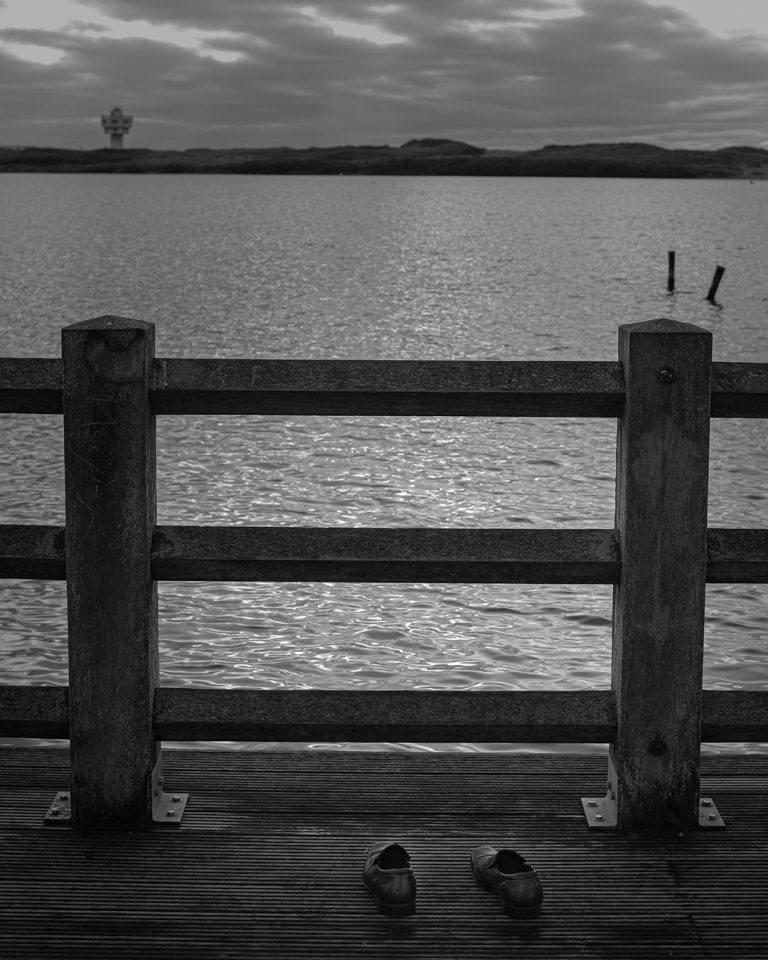 Black and white photograph of an empty pair of shoes left on a jetty overlooking water, conceptual photography project by Leon Britton